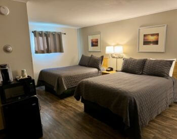 tan room with 2 double beds with brown quilts with stand in between with light above, wood floor, photos above beds