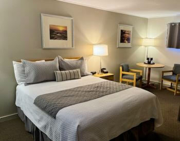 Tan Room with cream ceiling with a Queen sized bed, nightstand with lamp, round table in corner with 2 chairs, photos on wall