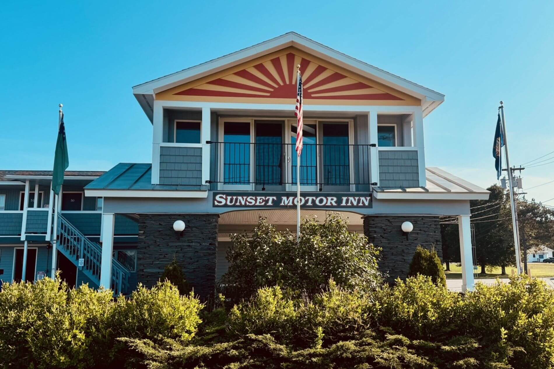 Gray 2-story building with sign "Sunset Motor Inn", orange and yellow streaks in form of sunshine above 2nd floor, flags on poles in front