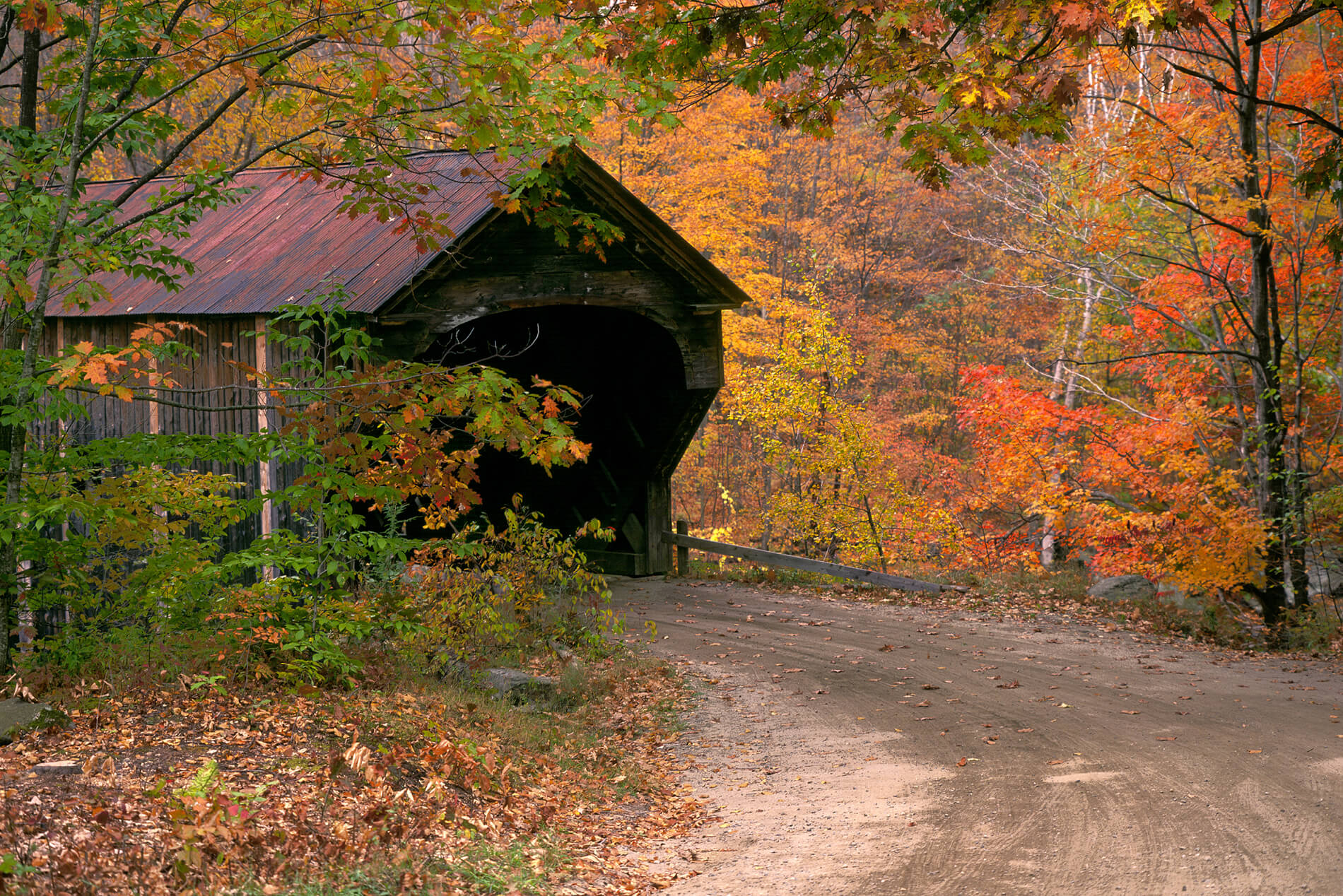 Rustic covered bridge along a dirt road in autumn with orange, green, and yellow leaves.