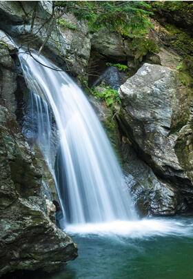 small waterfall landing into a swimming hole with green moss covered gray rocks.